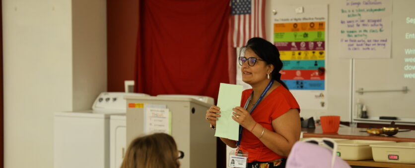 A teacher holds up a piece of paper while addressing her classroom