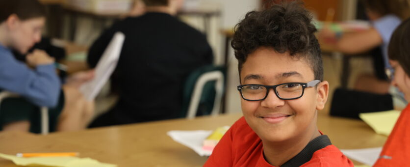 A middle school boy smiles while sitting at a table in a classroom