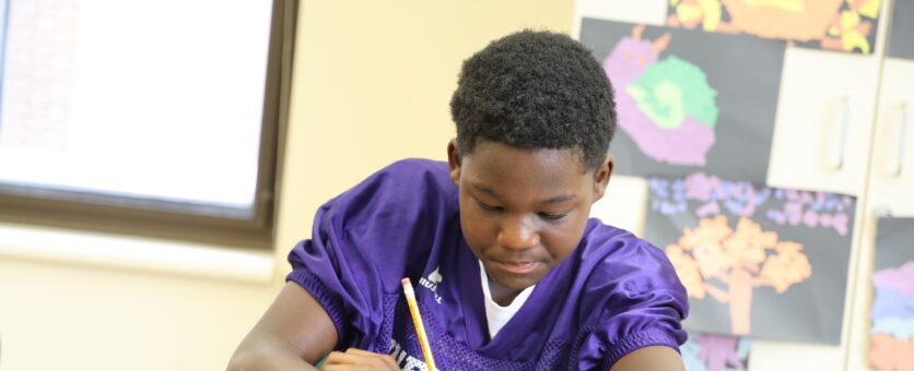 A middle school boy writes on a piece of paper while completing an assignment at his table.