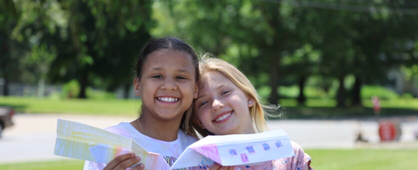 Two girls smile while standing outside and holding up the paper airplanes they built