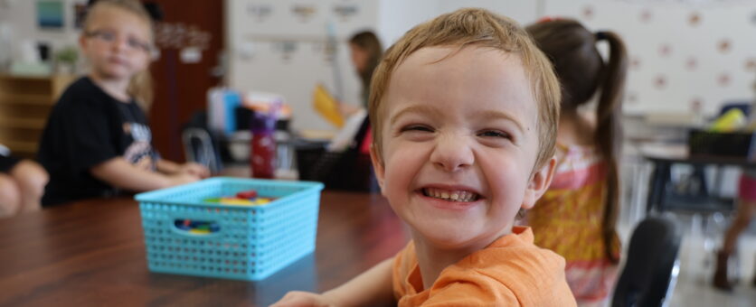 A young child shows off a huge grin while sitting at a table in his classroom on the first day of kindergarten.