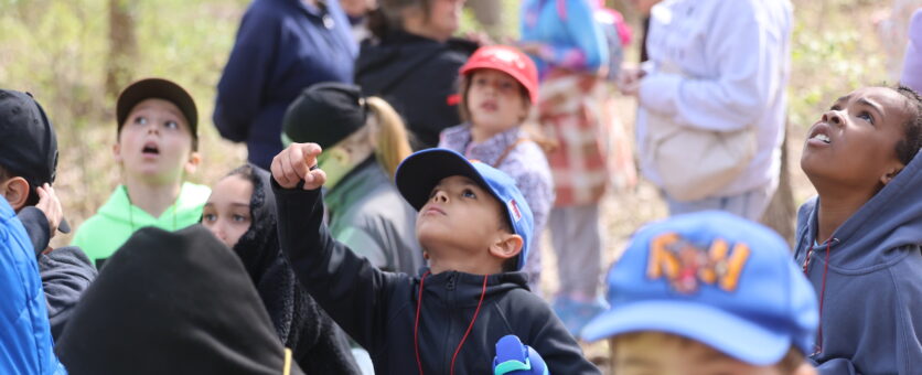 A boy holding a compass points up at the sky while on an outdoor field trip.