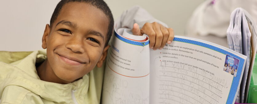 A boy proudly holds up a writing workbook he has been working in.