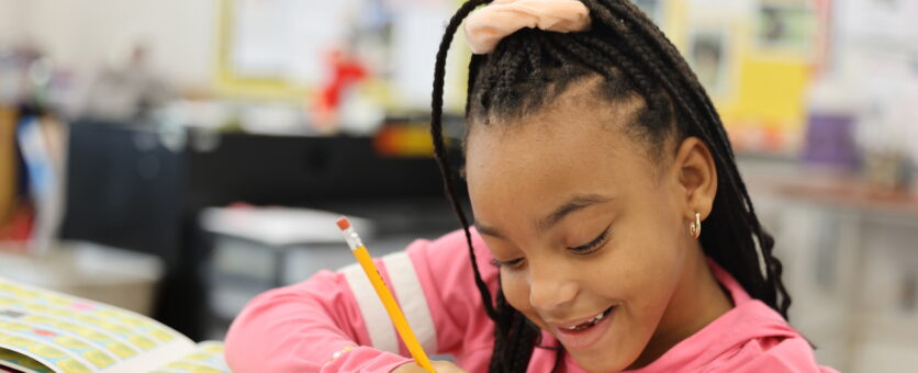 A girl smiles while using a pencil to write on a piece of paper at her desk in her classroom.