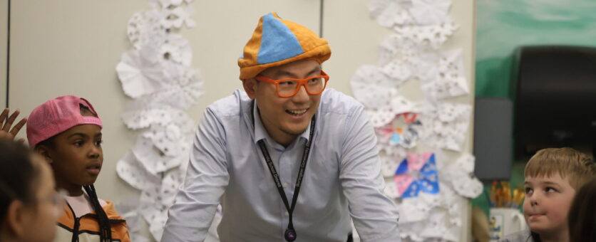 A teacher wearing a fun hat smiles while standing between two rows of students in his art class.