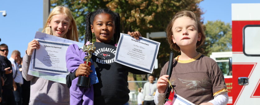 Two girls and a boy hold up certificates they won for their fire prevention posters while standing outside