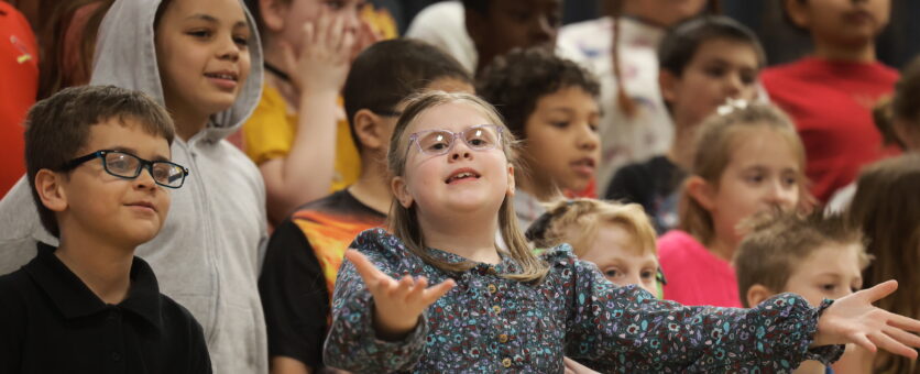 A young girl raises her arms while singing with a group of classmates during a concert