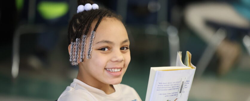 A girl smiles while reading a book in her classroom at Grimes Elementary School