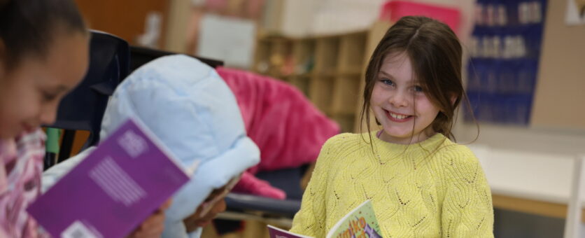 A girl smiles while reading a book in her classroom at Grimes Elementary School