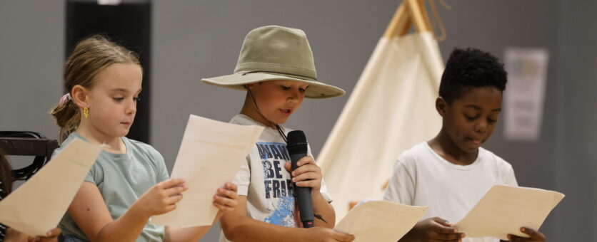 Two boys and a girl stand before their peers during an assembly in the school gym. The boy in the middle wears a hat and holds a microphone.