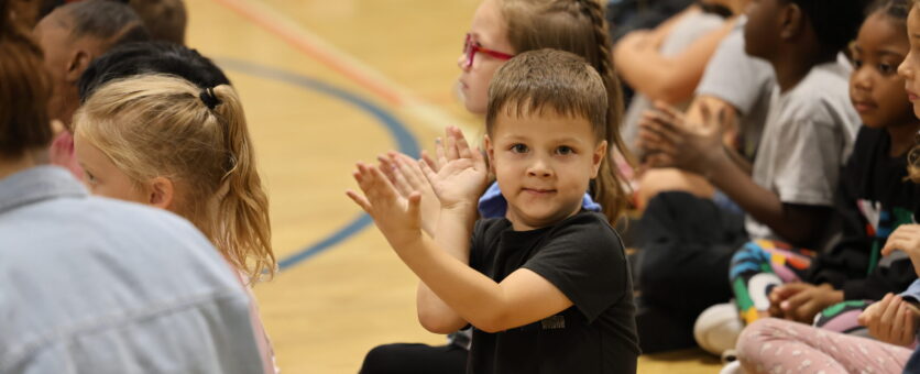 A young student looks at the camera while clapping his hands during an assembly about reading.